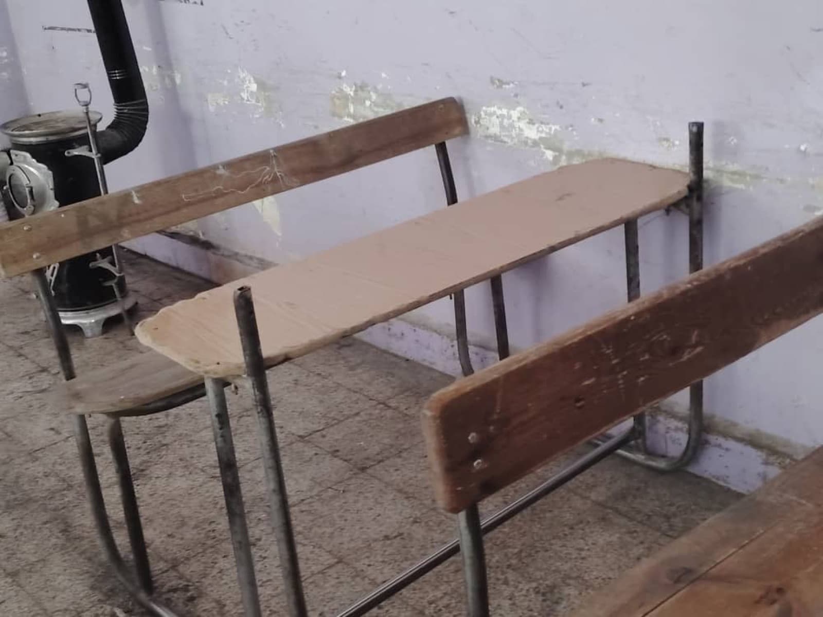 Abgenutzte Holzschulbänke neben einem alten Ofen in einem kahlen Klassenzimmer